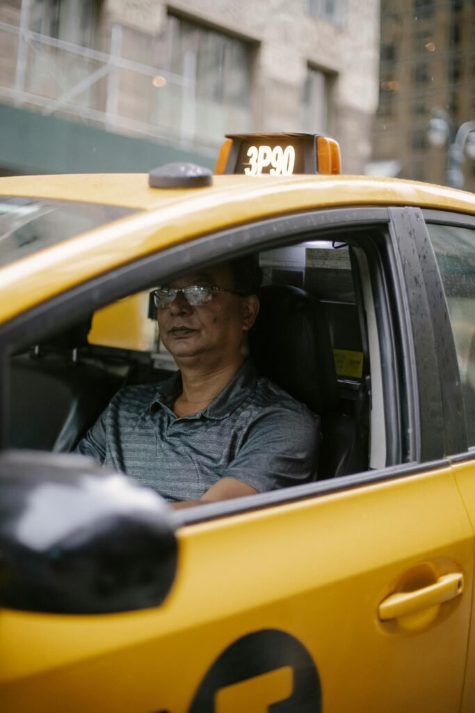 Serious middle aged ethnic man in polo shirt and eyeglasses driving modern yellow taxi car on city street in daytime